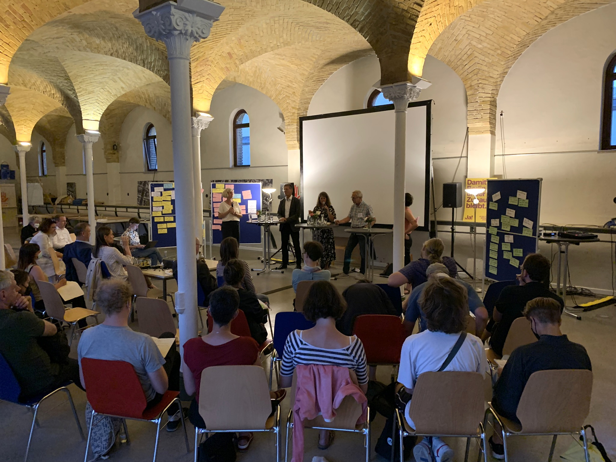 Rund 20 Personen sitzen in einem Halbkreis und blicken auf ein Podium mit vier Personen.